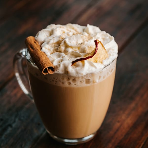 A clear mug of creamy beverage topped with whipped cream, a cinnamon stick, and a dried apple slice.
