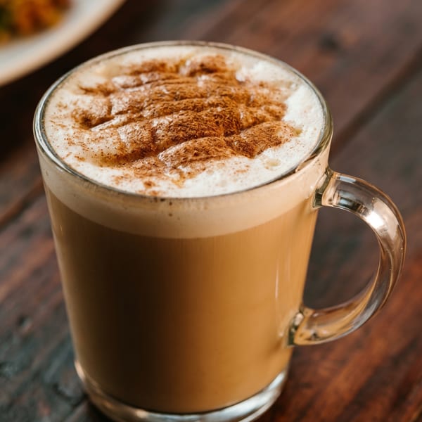 A clear mug of latte with foamed milk and cinnamon powder on a dark wooden table.